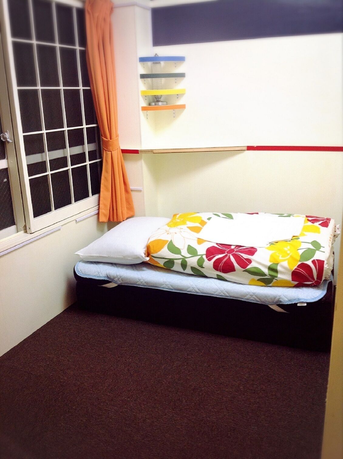 A small room with a window, a bed with floral bedding, a curtain, and a shelf with colorful items.