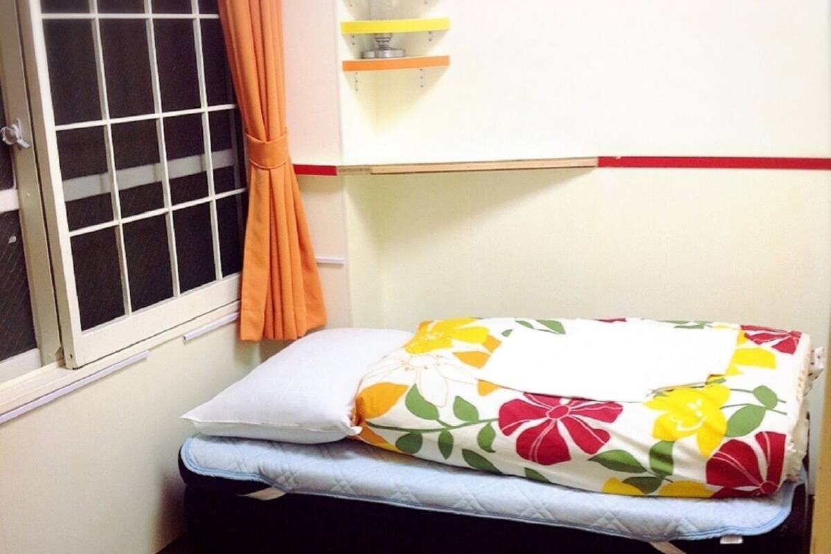 A small room with a window, a bed with floral bedding, a curtain, and a shelf with colorful items.