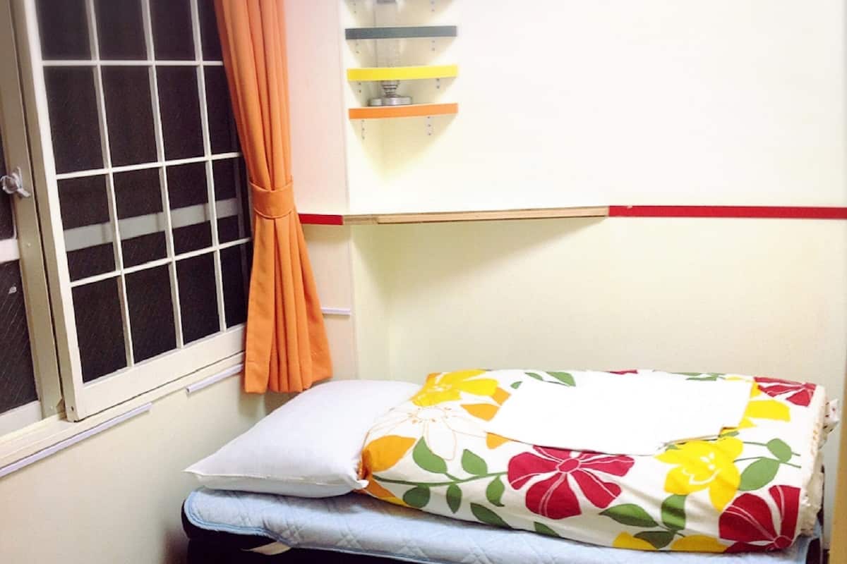 Ein kleines Zimmer mit Fenster, einem Bett mit floralem Überwurf, einem Vorhang und einem Regal mit Gegenständen.