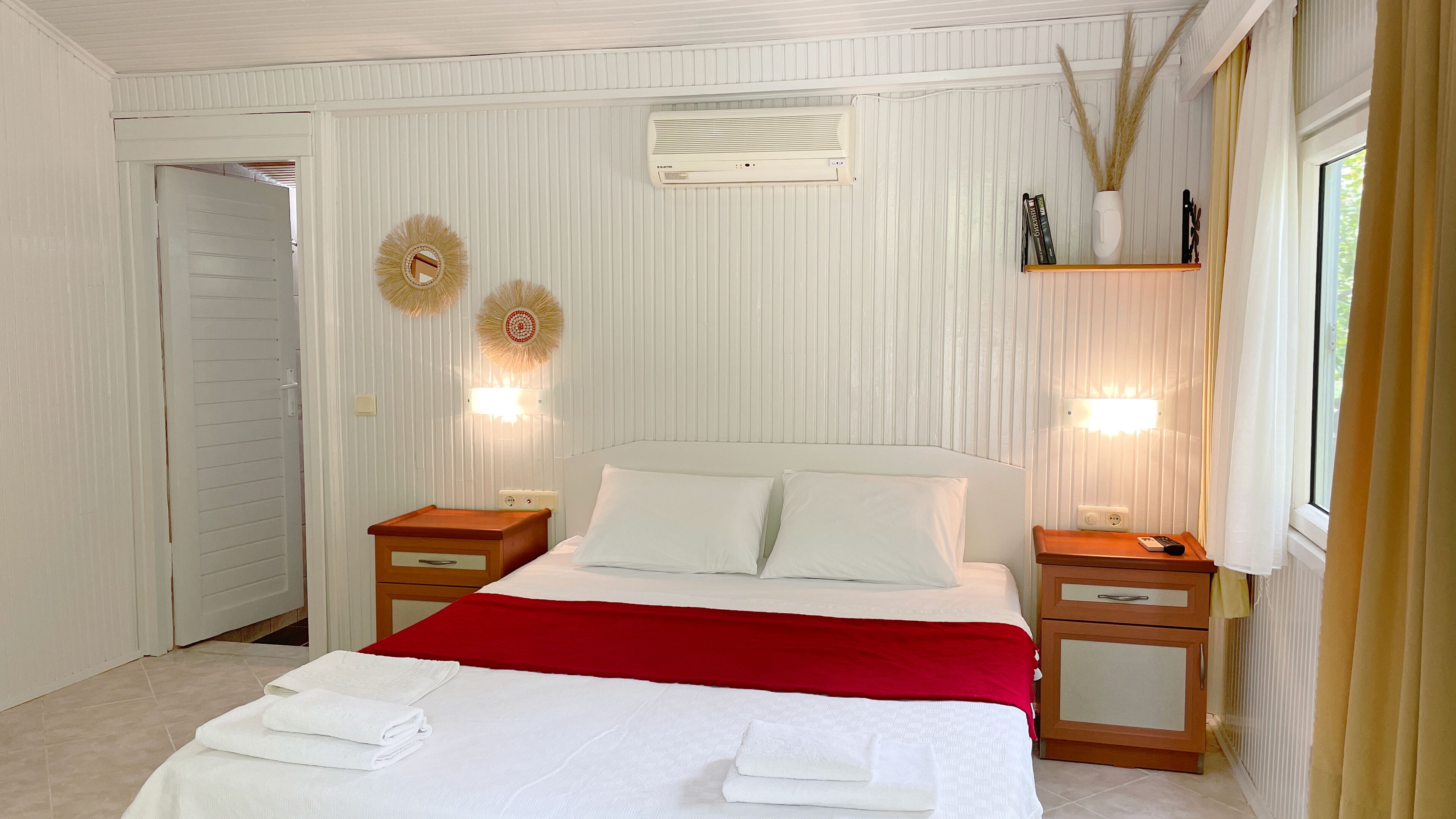 Una cama bien hecha con un edredón rojo, dos mesitas de noche con lámparas y un aire acondicionado de pared.