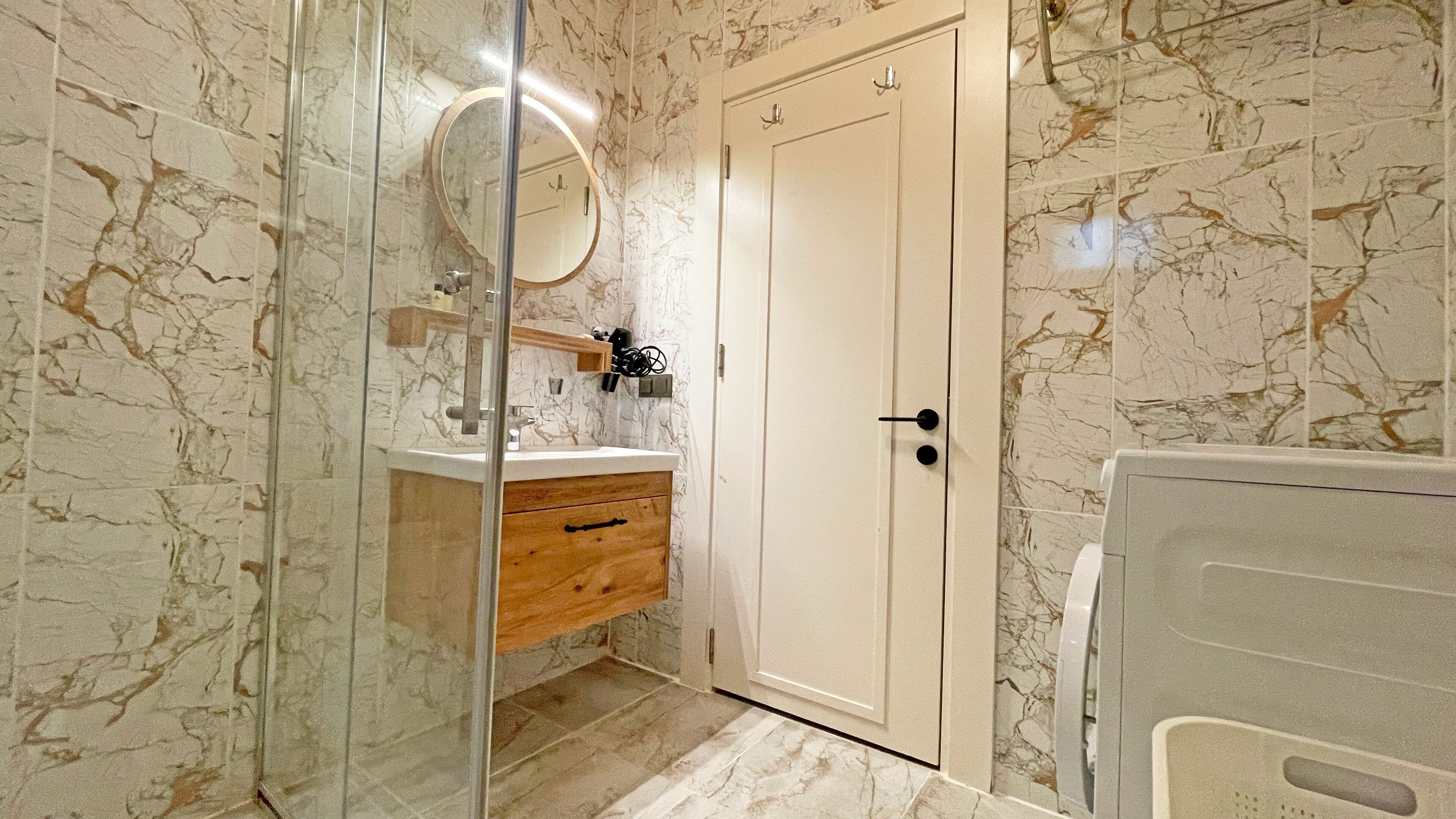 A bathroom with marble walls, a wooden vanity, a glass shower enclosure, and a white door.