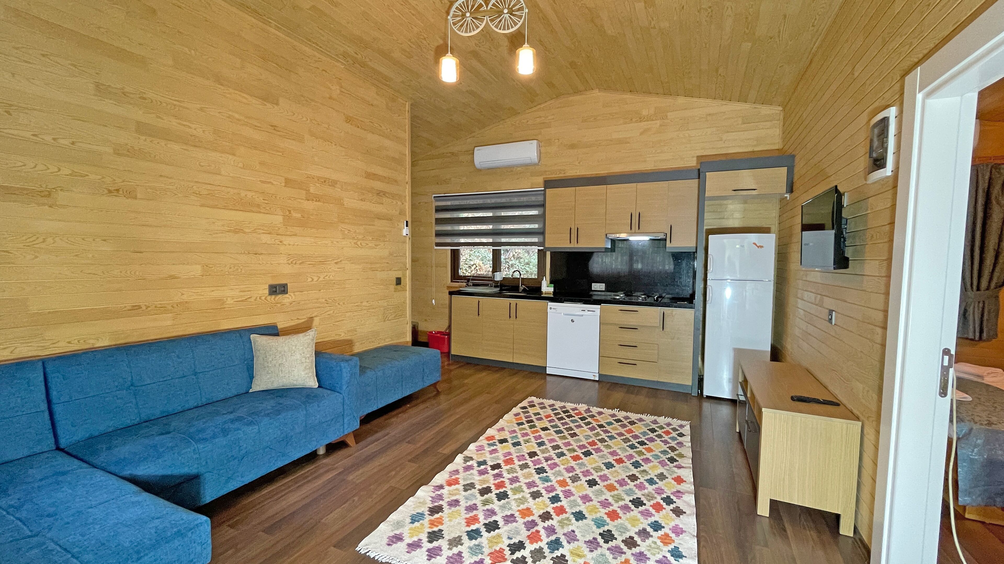 Um espaço de estar compacto com um sofá azul, uma cozinha com eletrodomésticos modernos e teto de madeira.