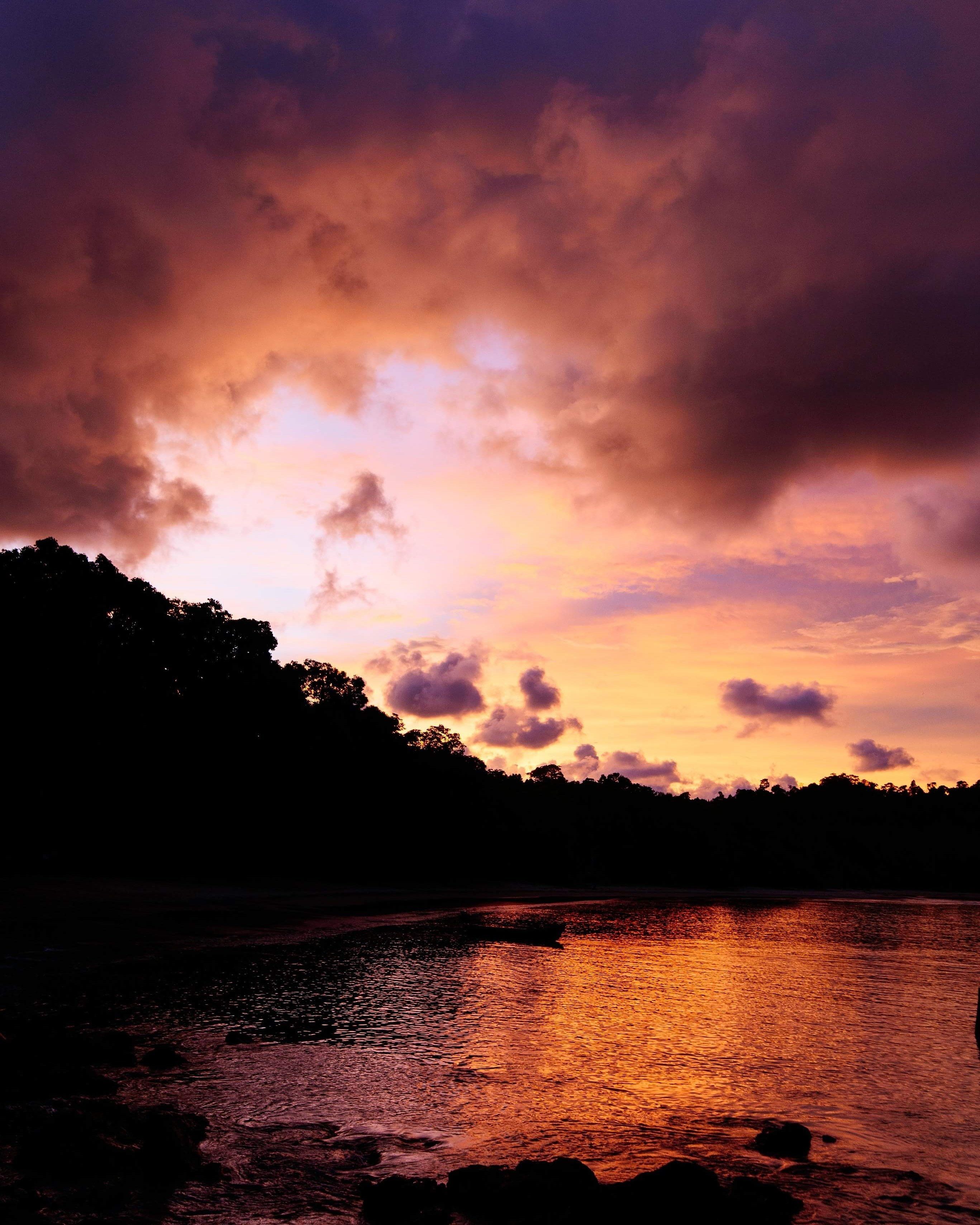 A sunset over a body of water with a silhouette of trees and a rocky shoreline.