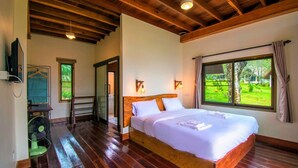 Une chambre spacieuse avec un grand lit, un plancher en bois, une vue sur la verdure par la fenêtre et un ventilateur au sol.