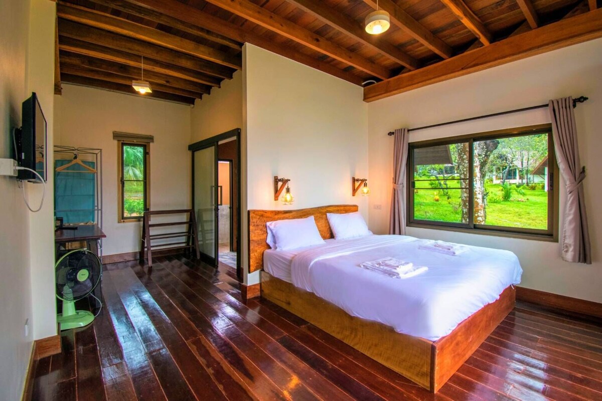 Un'ampia camera da letto con un letto grande, pavimento in legno, vista sul verde dalla finestra e una ventola a terra.