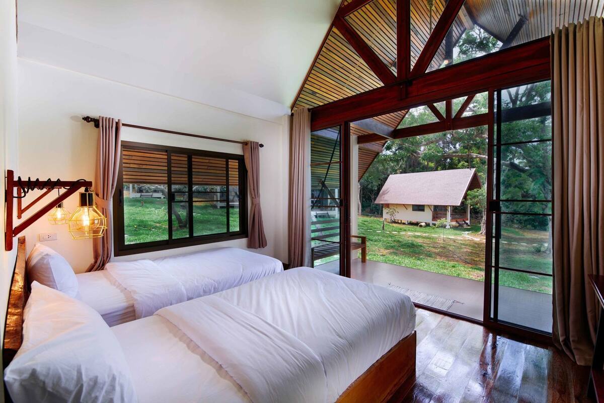 A bedroom with two beds, a large window, and a view of a house.