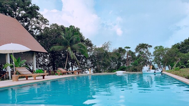 Une piscine entourée d’arbres et d’un bâtiment à toit de tuiles, avec des chaises longues et des parasols à proximité.