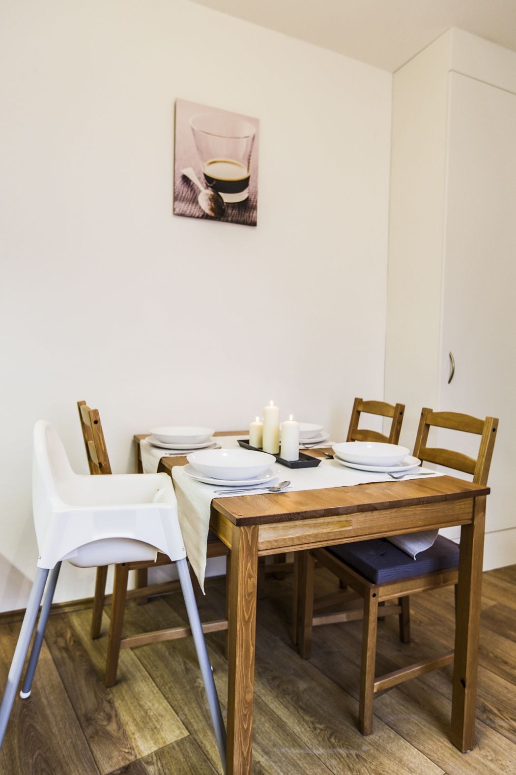 A dining area with a wooden table, white chairs, and a small white tablecloth. There are candles on the table, and a framed picture hangs on the wall above.