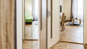 Een gang met houten vloeren, een spiegelkast en uitzicht op een kamer met een bed, een bureau en een stoel.