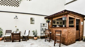 Een terras met stenen vloer, houten stoelen, een klein houten kioskje en potplanten.