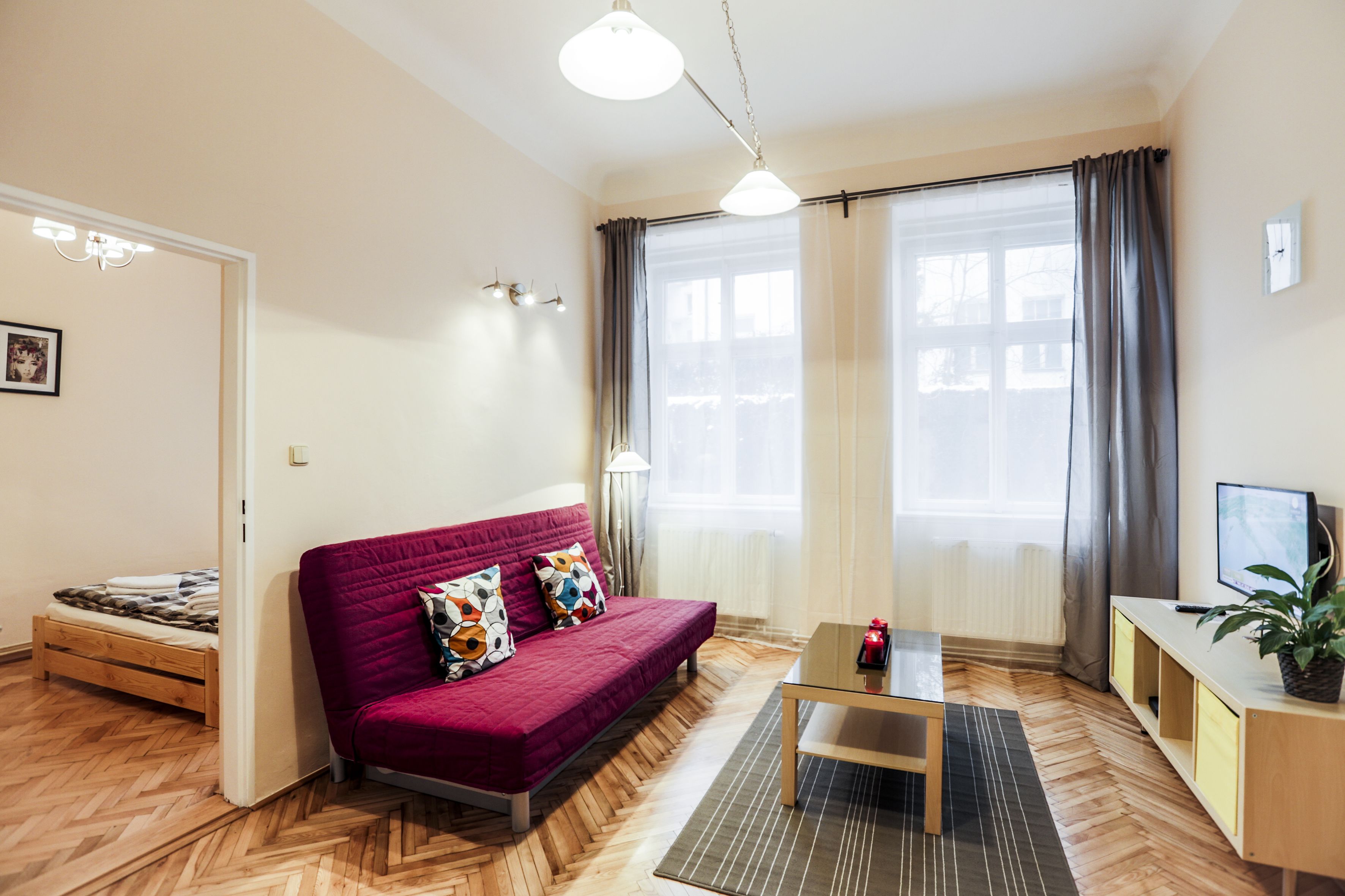 Ein modernes Wohnzimmer mit Holzboden, einem roten Sofa mit bunten Kissen, einem Glastisch und einem kleinen Holznachttisch, sowie einer Topfpflanze.