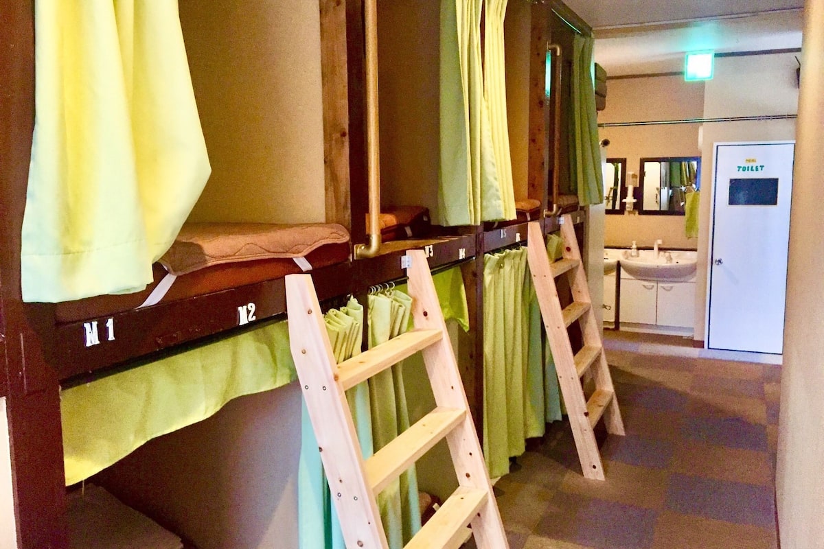 A row of bunk beds with green curtains, a wooden ladder, and a visible bathroom with a sink and mirror.