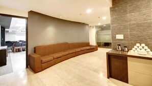 A modern reception area with a curved brown sofa, a wooden reception desk with coffee and cups, and a marble floor. - Hotel Good Times (New Delhi)