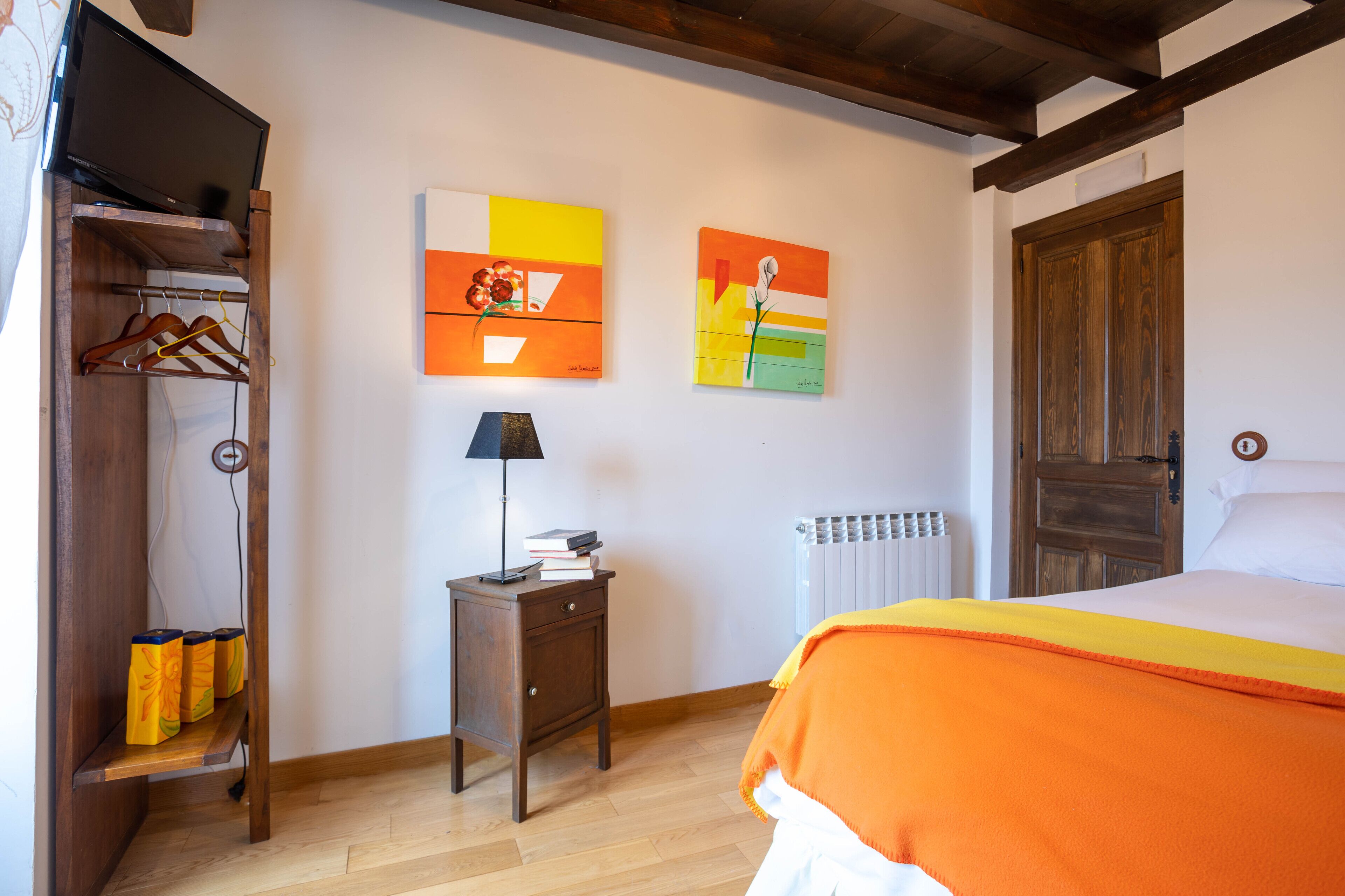 A bedroom with a wooden wardrobe, a bedside table with a lamp, and two paintings on the wall.
