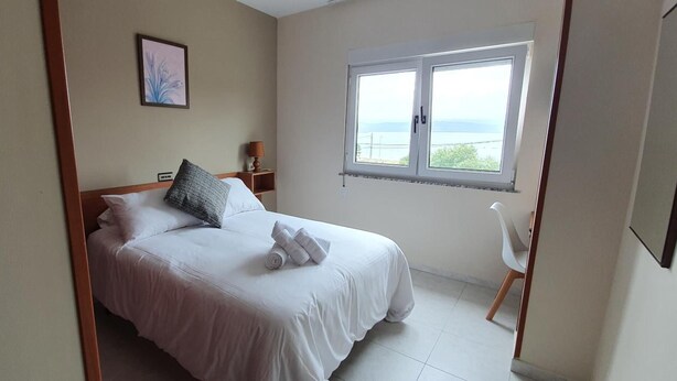 A hotel room with a bed, a window with a view, a bedside table with a lamp, and a chair. - Hotel Arenal (Fisterra)