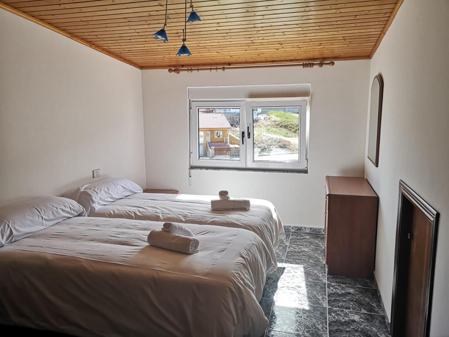 A bedroom with two beds, a window with a view, a wooden ceiling, and a marble floor.
