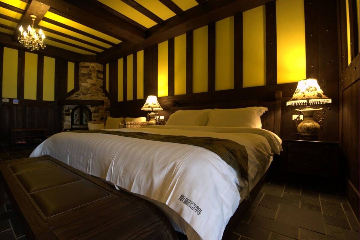 A room with a large bed, wooden beams, stone fireplace, and two wall-mounted lamps.
