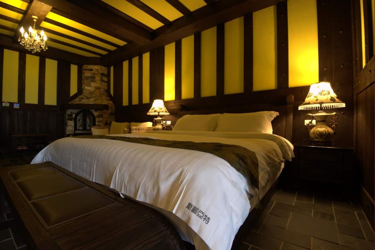 A hotel room with a large bed, two bedside lamps, a stone fireplace, and a wooden bench.
