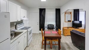 A compact kitchen with white cabinets, a stove, and a sink. There is a dining table with chairs and a television on a wooden dresser. - Richards Motel Courtyard (Hollywood)
