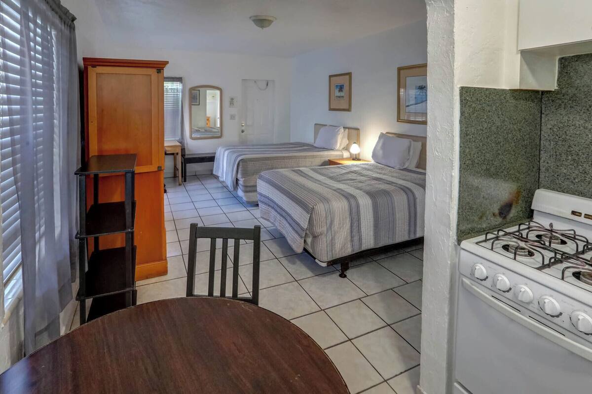 A compact room with two beds, a wooden chair, a dining table, and a kitchen area with a stove.