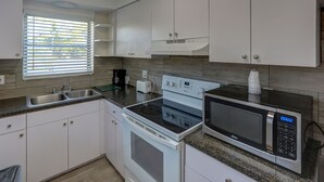 A modern kitchen with white cabinets, a built-in oven, a microwave, and a window with blinds. - Richards Motel Courtyard (Hollywood)