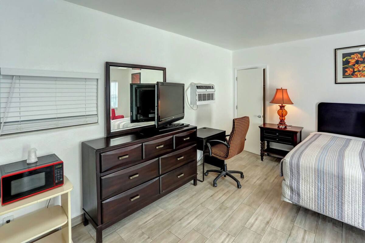 A hotel room with a bed, a desk with a computer and monitor, a chair, a microwave, a lamp, and a framed picture on the wall.