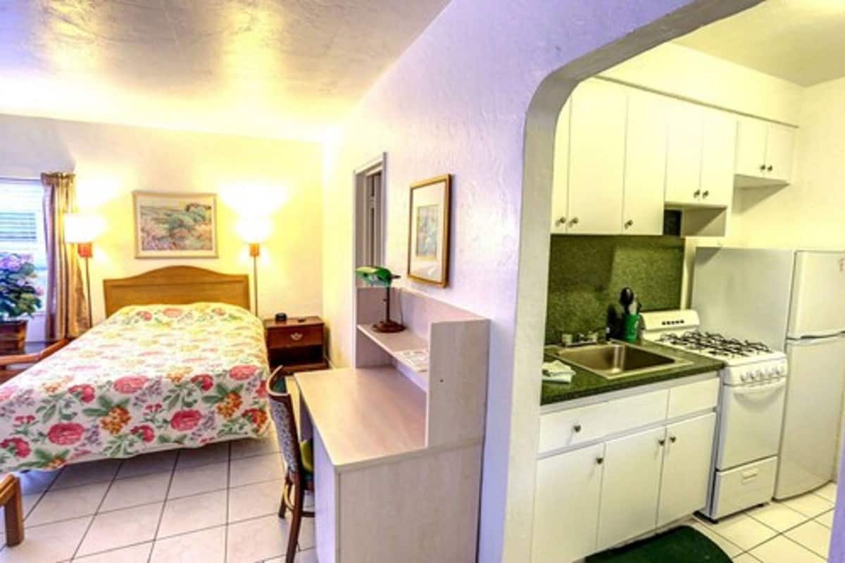 A compact hotel room with a bed, bedside table, lamp, and a kitchenette with a stove, sink, and cabinets.