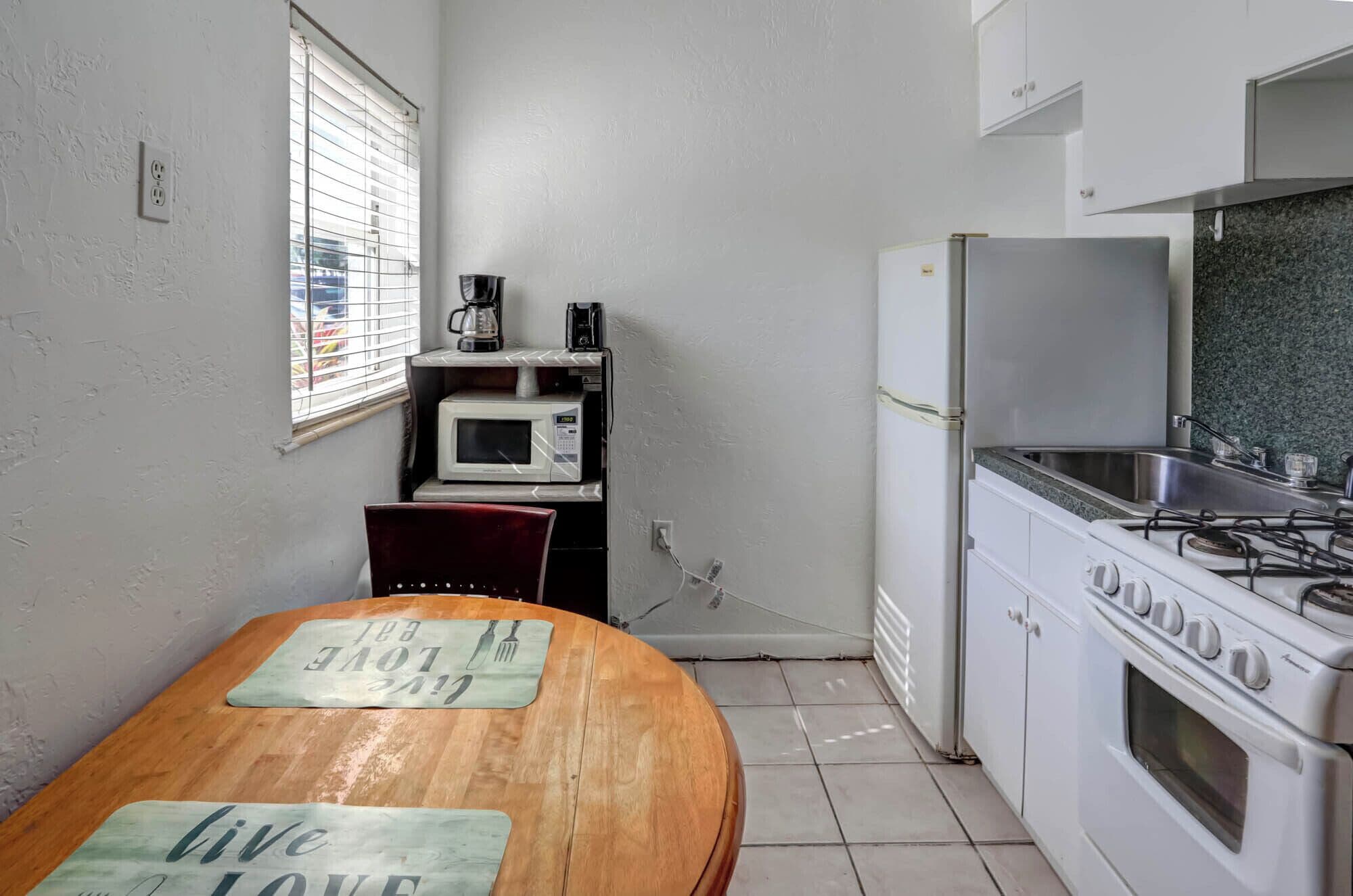 Cocina compacta con estufa, microondas y refrigerador. Hay una ventana con persianas, una mesa de madera con individuales y piso de baldosas.