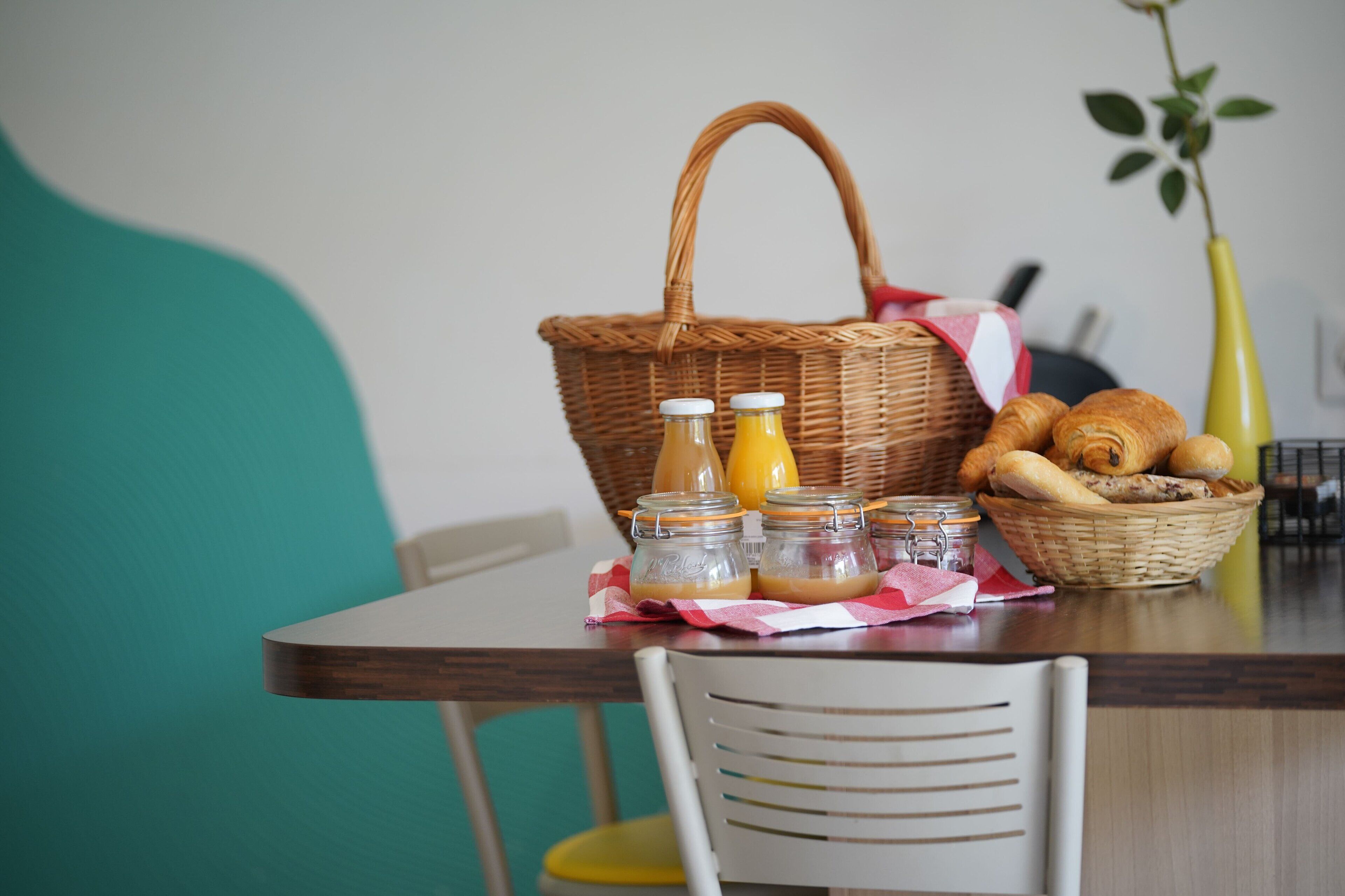 Une table sur laquelle se trouve un panier en osier contenant du pain, un pot de confiture et une bouteille de jus de fruits.