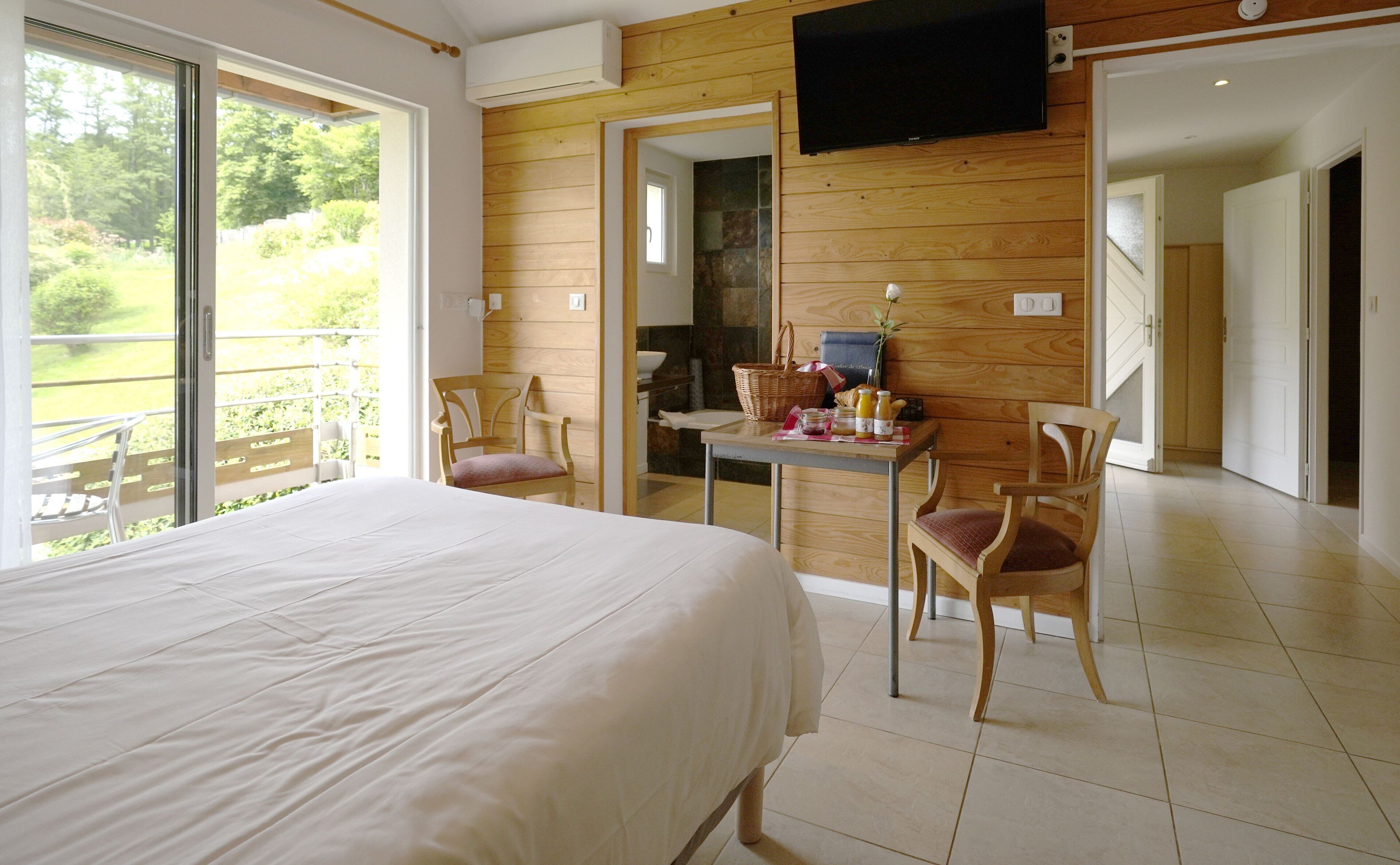 Een slaapkamer met een groot bed, een eettafel met stoelen, een aan de muur bevestigde televisie en een balkon met uitzicht op groen.