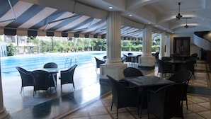 A covered outdoor seating area with tables and chairs, a pool in the background, and a view of the surrounding greenery. - The Classique Club (Mumbai)