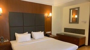 A hotel room with a large bed, wooden headboard, bedside tables, a nightstand, and a mirror on the wall. - The Classique Club (Mumbai)