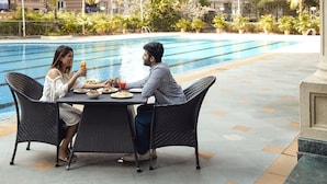 A dining area with a pool view, outdoor seating, and a modern building in the background. - The Classique Club (Mumbai)