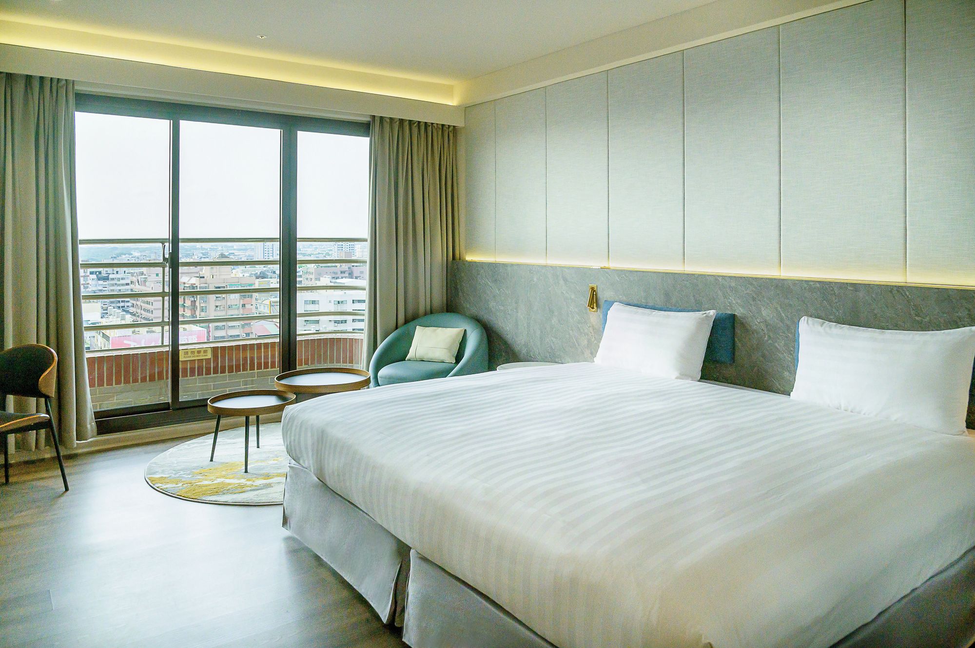 A hotel room with a large bed, a seating area with a chair and a small table, and a view of the city through large windows.