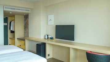 A hotel room with a bed, a desk with a flat-screen TV, a small refrigerator, a clock, and a wardrobe.