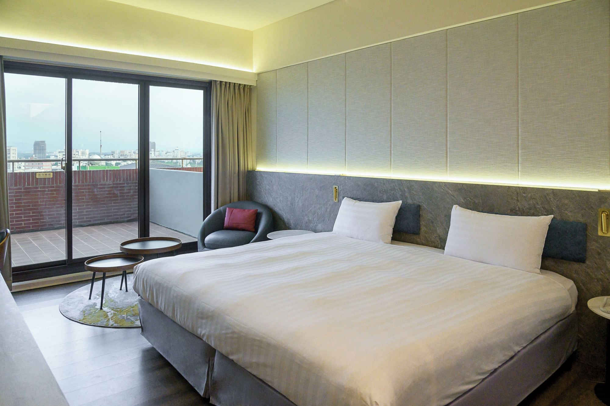 A hotel room with a large bed, a chair, a small table, and a view of the city through a balcony.