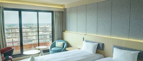 Habitación de hotel con un ventanal grande, dos camas, una silla, una mesita y vistas a la ciudad.