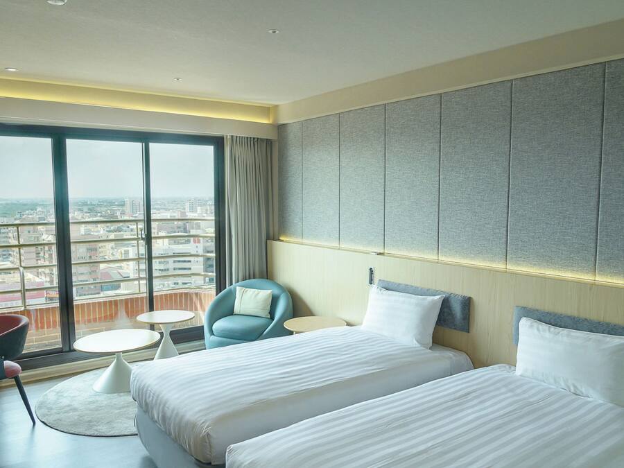 Ein Hotelzimmer mit großem Fenster, zwei Betten, einem Sessel, einem kleinen Tisch und Blick auf die Stadt.