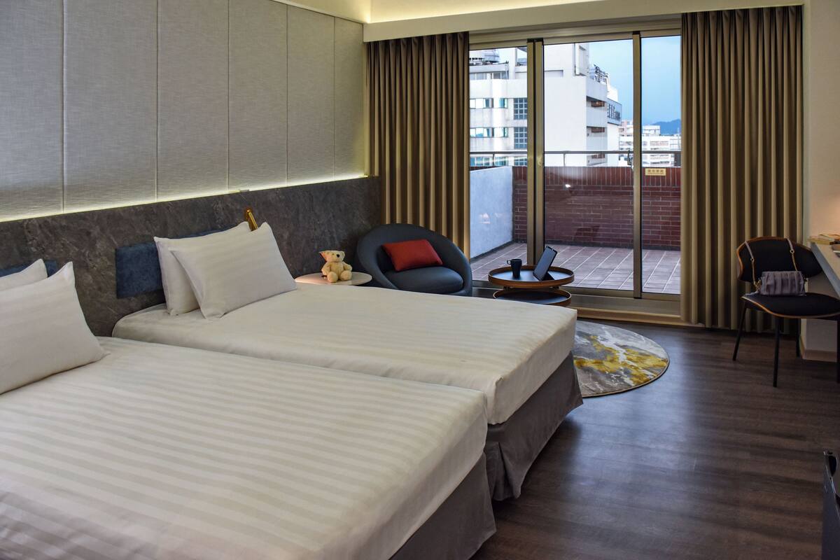 A hotel room with two beds, a desk, a chair, a teddy bear, and a view of the city through large windows.