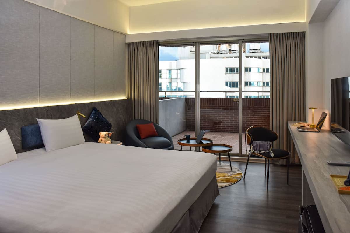 A modern hotel room with a large bed, two chairs, a small table, and a desk with a laptop.