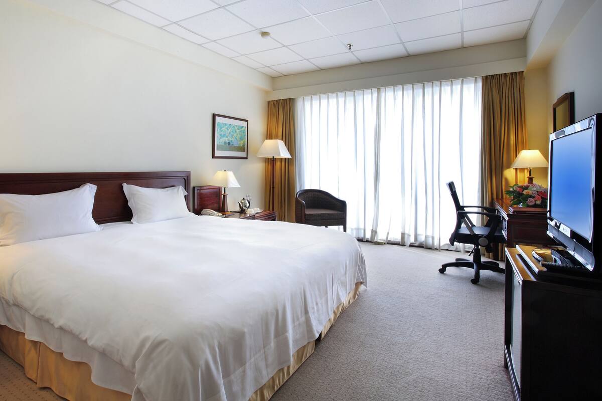 A hotel room with a large bed, a desk with a TV, a chair, a lamp, and a window with curtains.