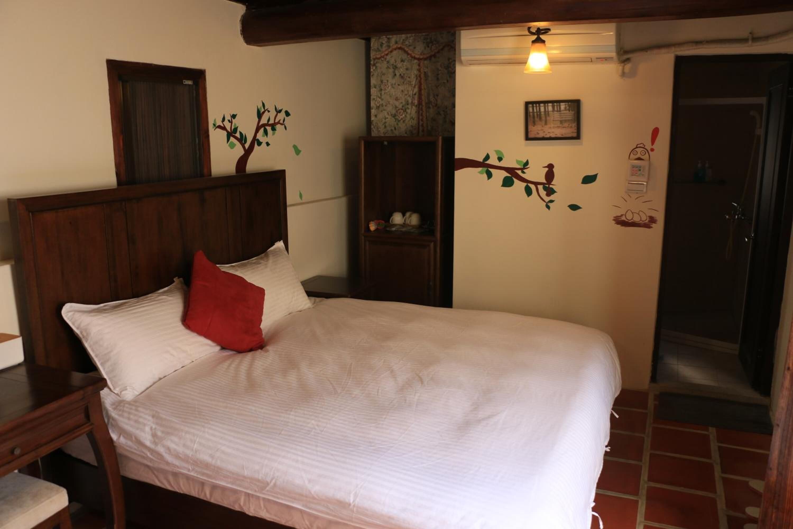 Un dormitorio con cama de madera, ropa de cama blanca, un almohadón rojo, un estante en la pared y una puerta.