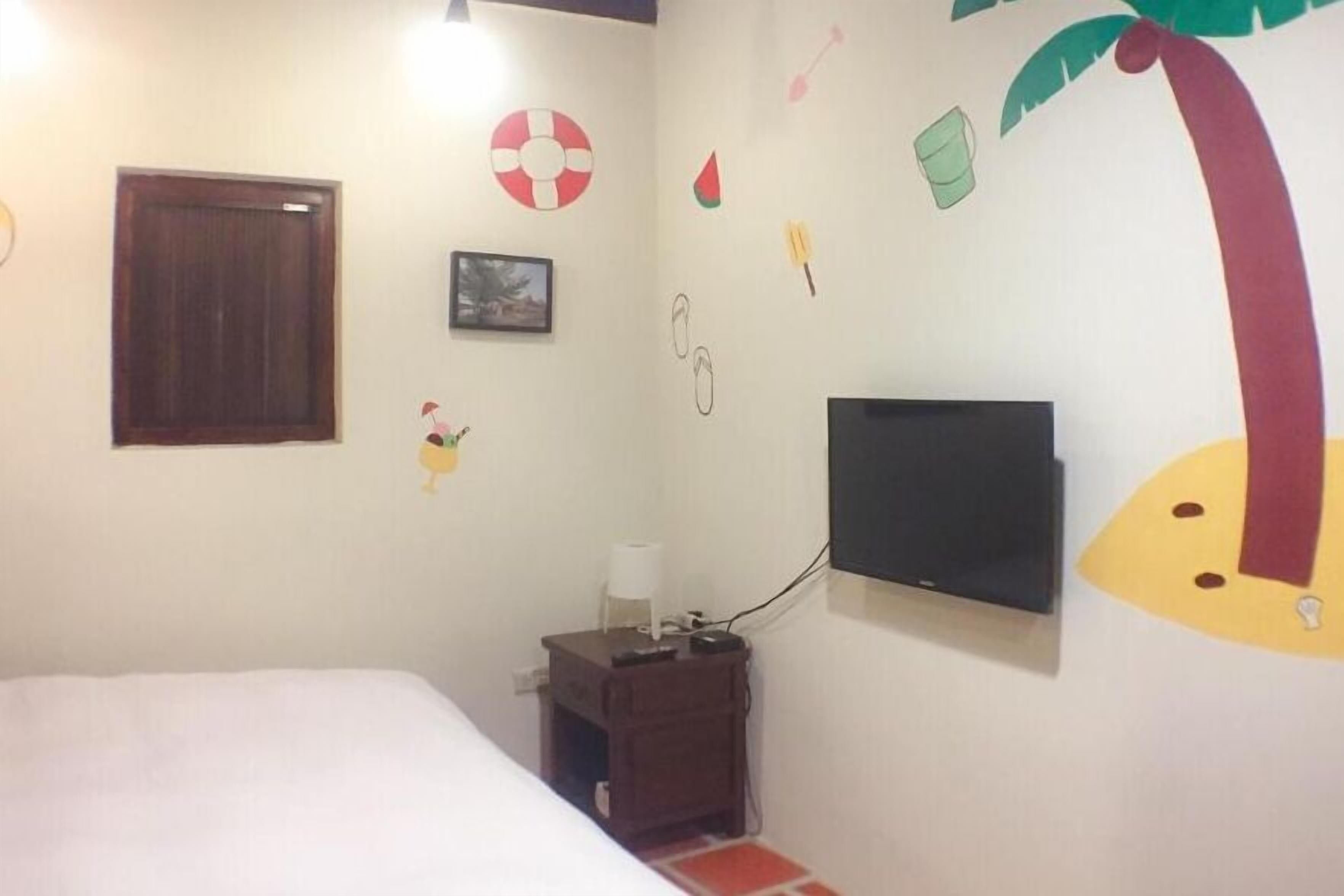 Une chambre à coucher comprenant un lit, un téléviseur fixé au mur, une petite table de chevet en bois et des éléments de décoration murale, notamment un palmier et une pastèque.