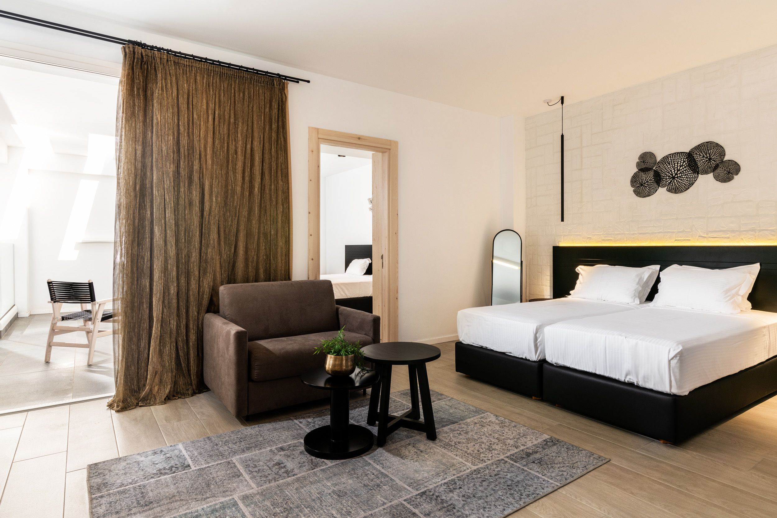 A modern hotel room with a bed, a sofa, a small table with a plant, and a large window with curtains.