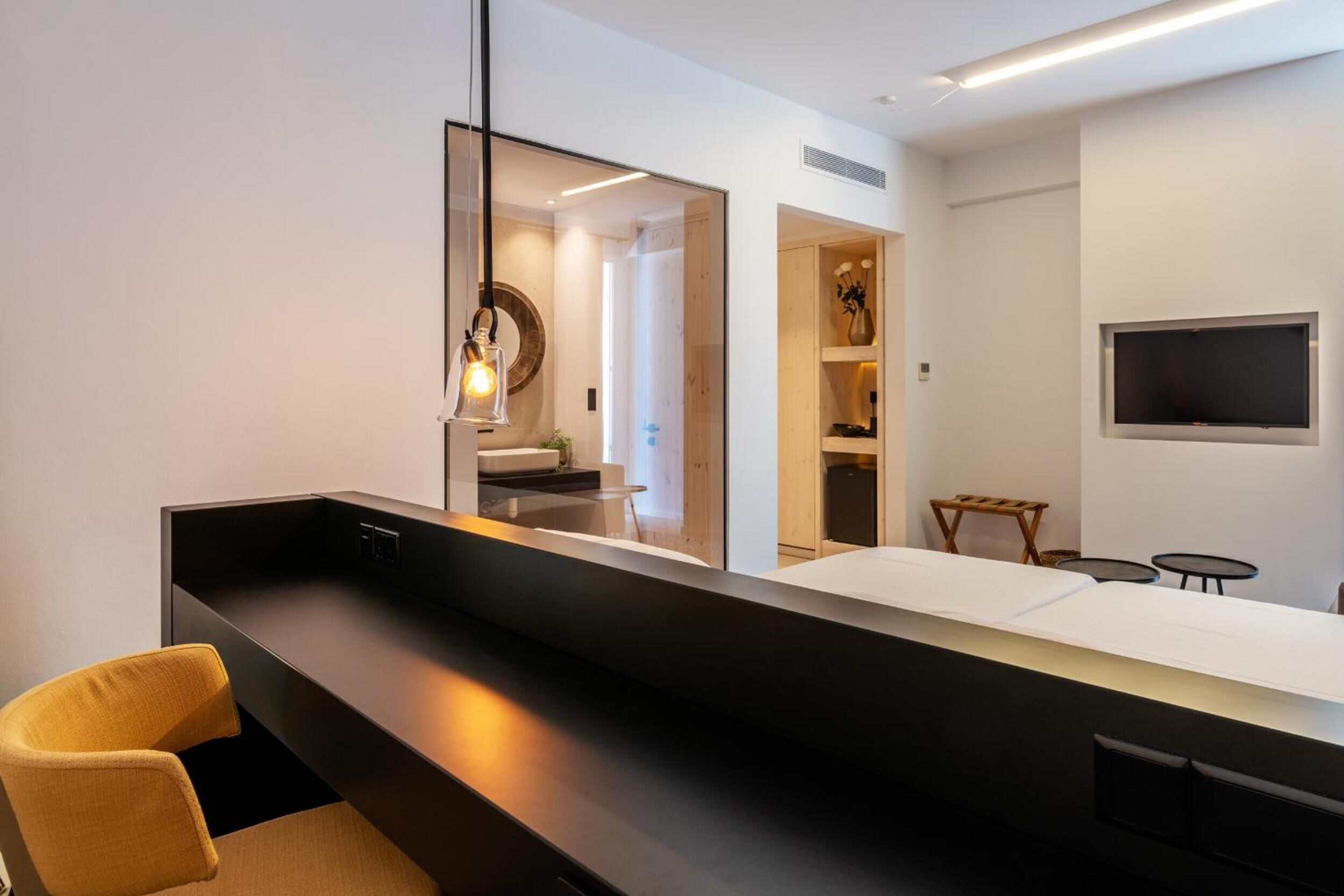 A modern hotel room with a long counter, a large mirror, a television mounted on the wall, and a small seating area with a chair and tables.