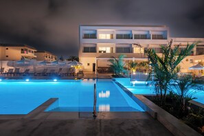 A swimming pool with lounge chairs and umbrellas, surrounded by a building with multiple balconies and a well-lit entrance.