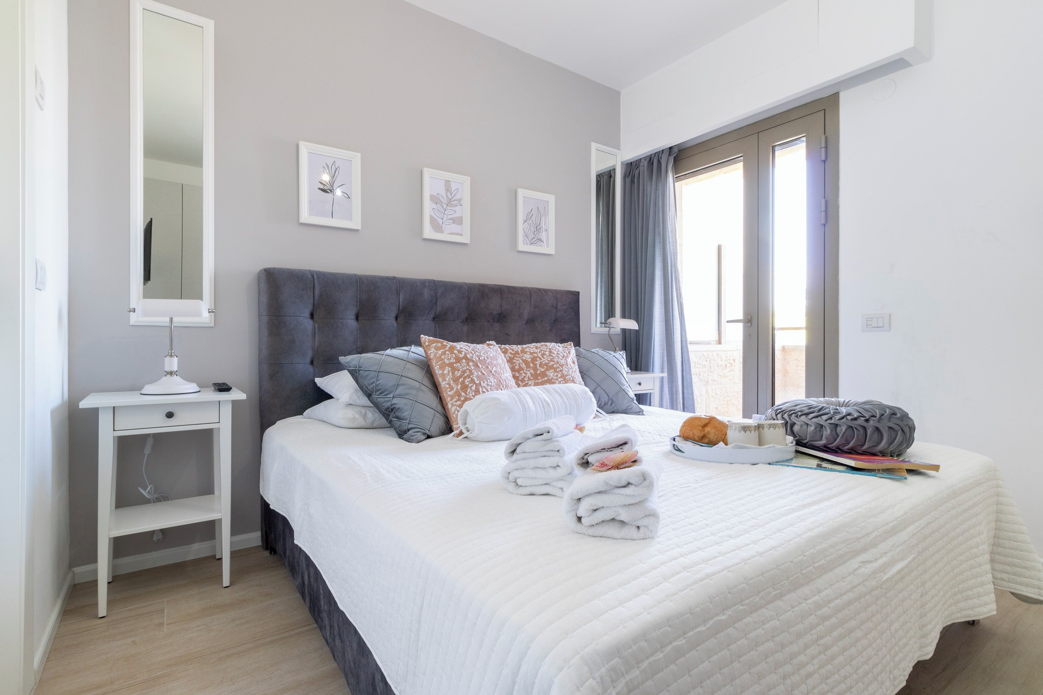 A neatly made bed with a grey headboard, white bedding, and a bedside table with a lamp.