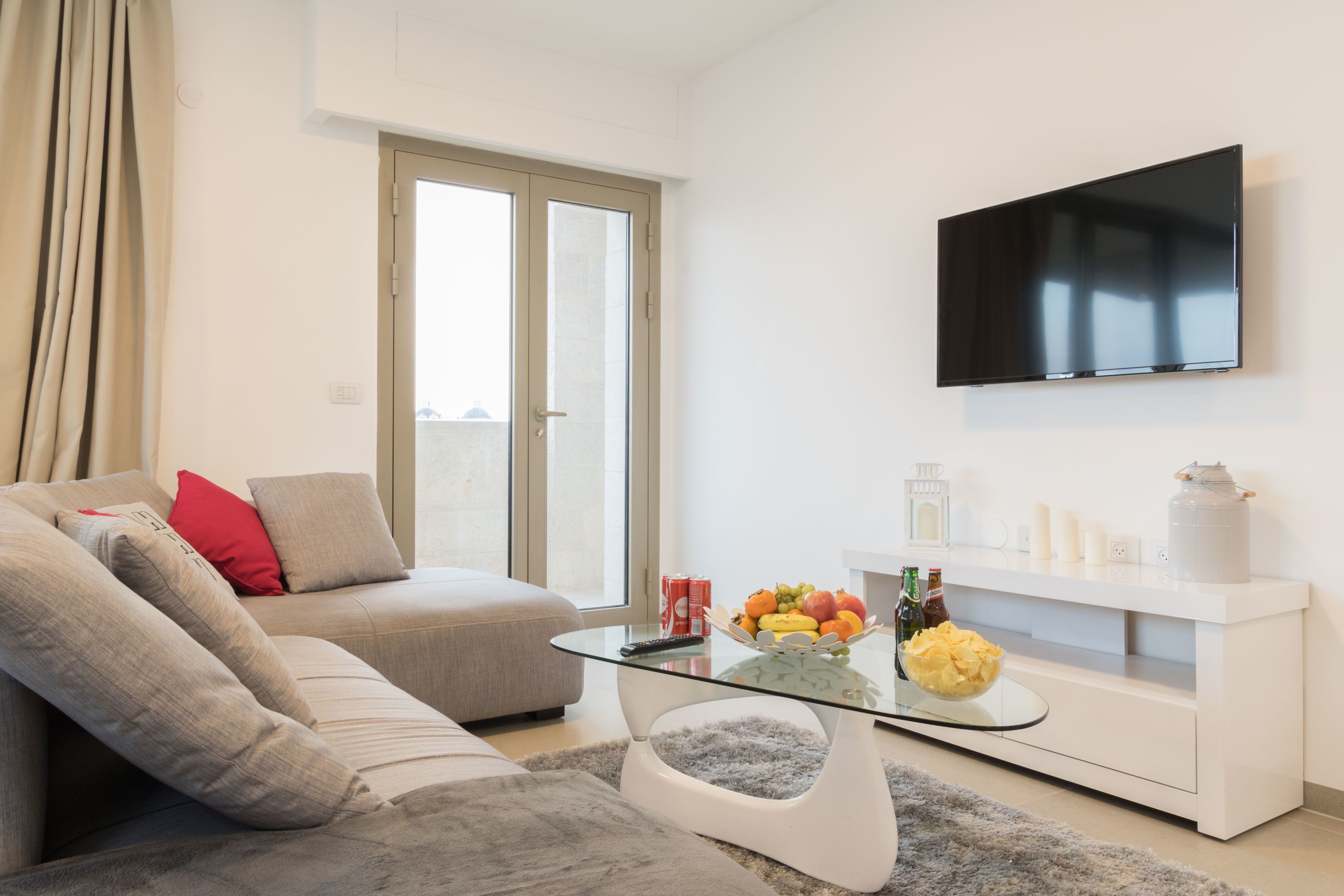 A modern living room with a glass coffee table, a sofa with cushions, a flat-screen TV mounted on the wall, and a small white cabinet with decorative items.