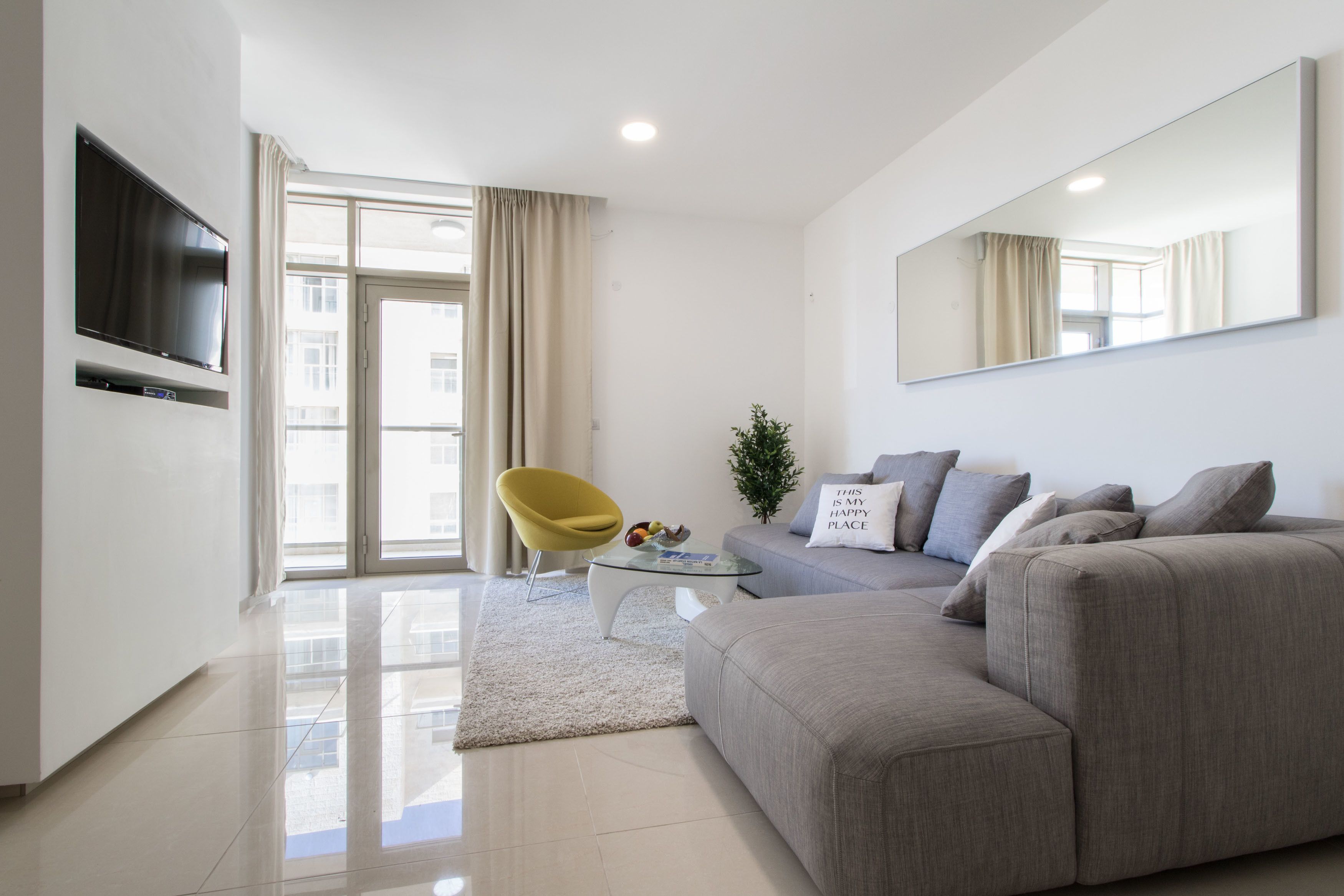 A modern living room with a grey sofa, a yellow chair, a glass coffee table, and a flat-screen TV mounted on the wall.