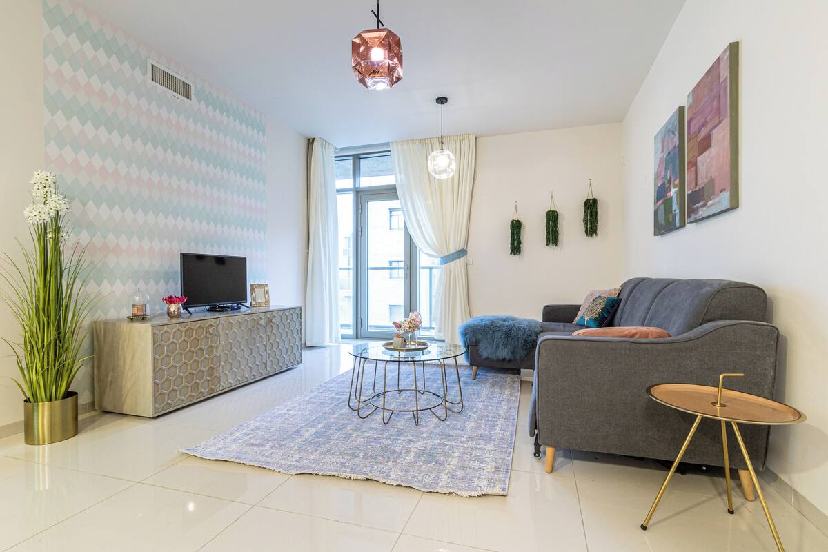 Ein modernes Wohnzimmer mit einem grauen Sofa, einem runden Couchtisch, einem Fernseher auf einer Anrichte und einem großen Fenster mit Vorhängen.